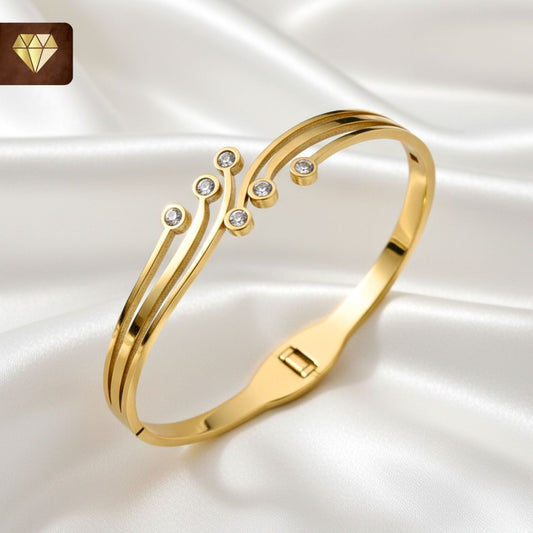 A close-up shot of a modern, gold-plated Kada bangle. The bracelet features a fluid, multi-band design with brilliant cubic zirconia stones placed along the curves. The finish is a high-shine polished gold.
